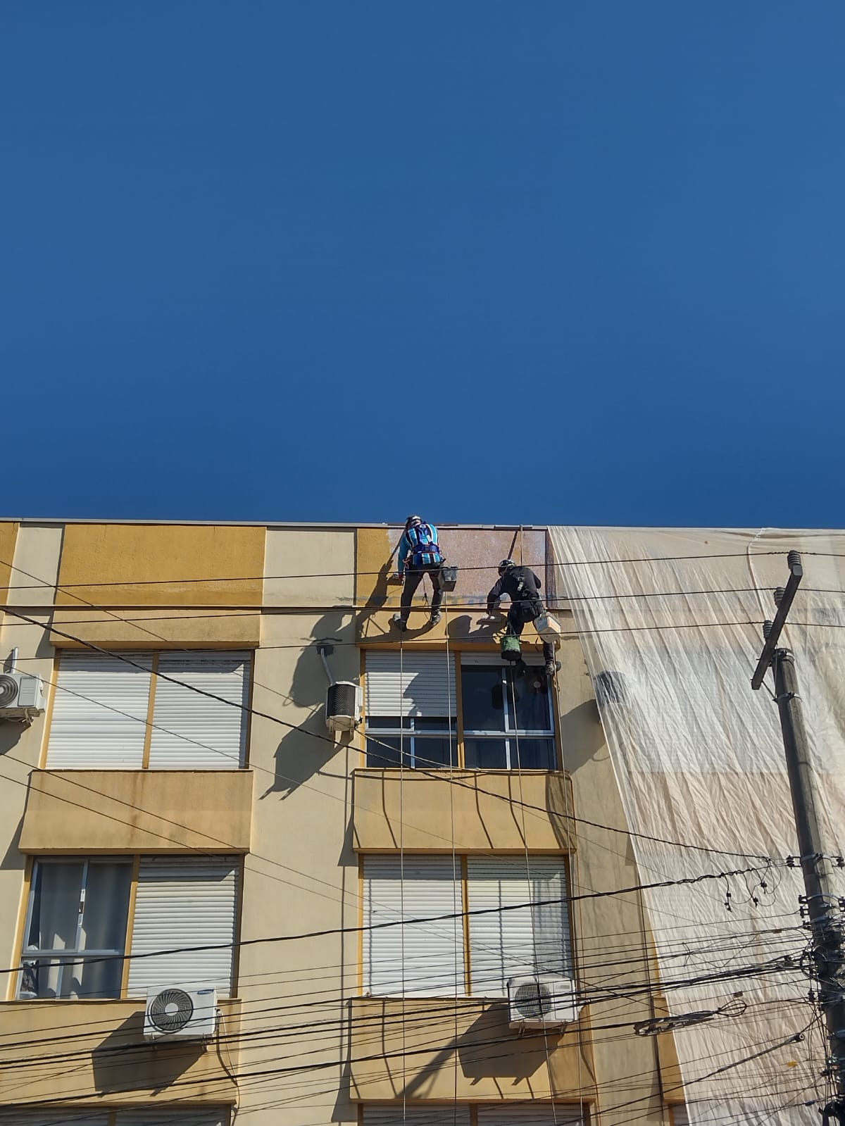 Dois alpinistas em um andaime suspenso, realizando manutenção em uma fachada.